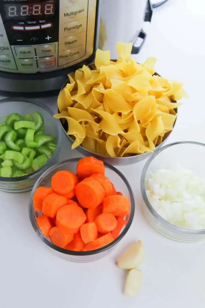 Chicken noodle soup ingredients sitting in individual bowls in front of an Instant Pot. Ingredients include egg noodles, garlic cloves, diced celery and carrots and chopped onions.
