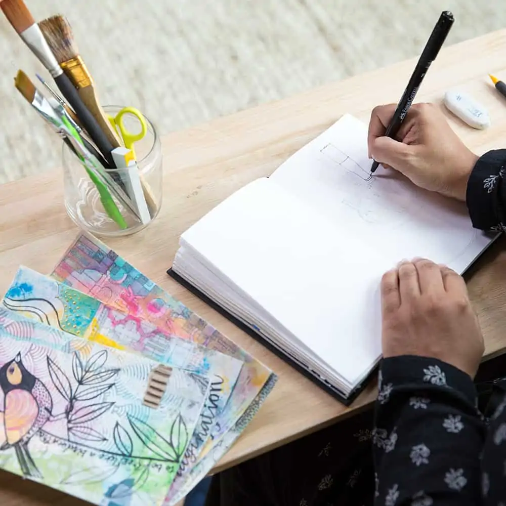 Woman doodling in notebook with paints, pens, and water colors on her desk.