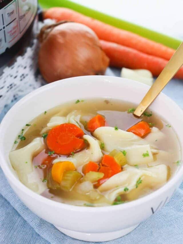 A bowl of chicken noodle soup with an onion and carrots sitting on a table beside it.