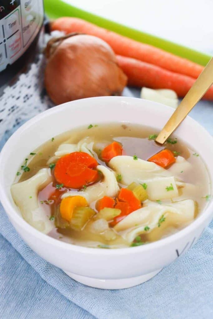 a bowl of chicken noodle soup with an onion and carrots sitting on a table beside it.
