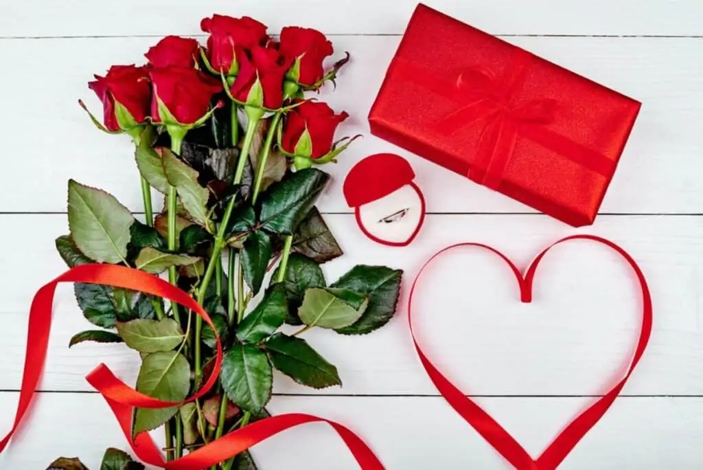 red roses sitting a table with a ring in a box and a wrapped gift for Valentine's Day.