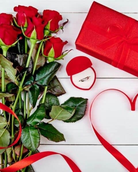 red roses sitting a table with a ring in a box and a wrapped gift for Valentine's Day.