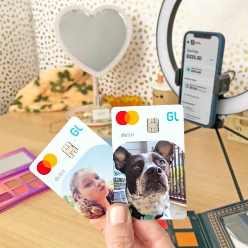 A hand holds two Greenlight debit cards with custom photos—one of a girl and one of a dog—in front of a phone displaying a banking app on a ring light stand.