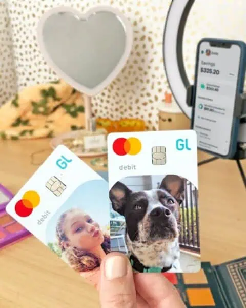 A hand holds two Greenlight debit cards with custom photos—one of a girl and one of a dog—in front of a phone displaying a banking app on a ring light stand.