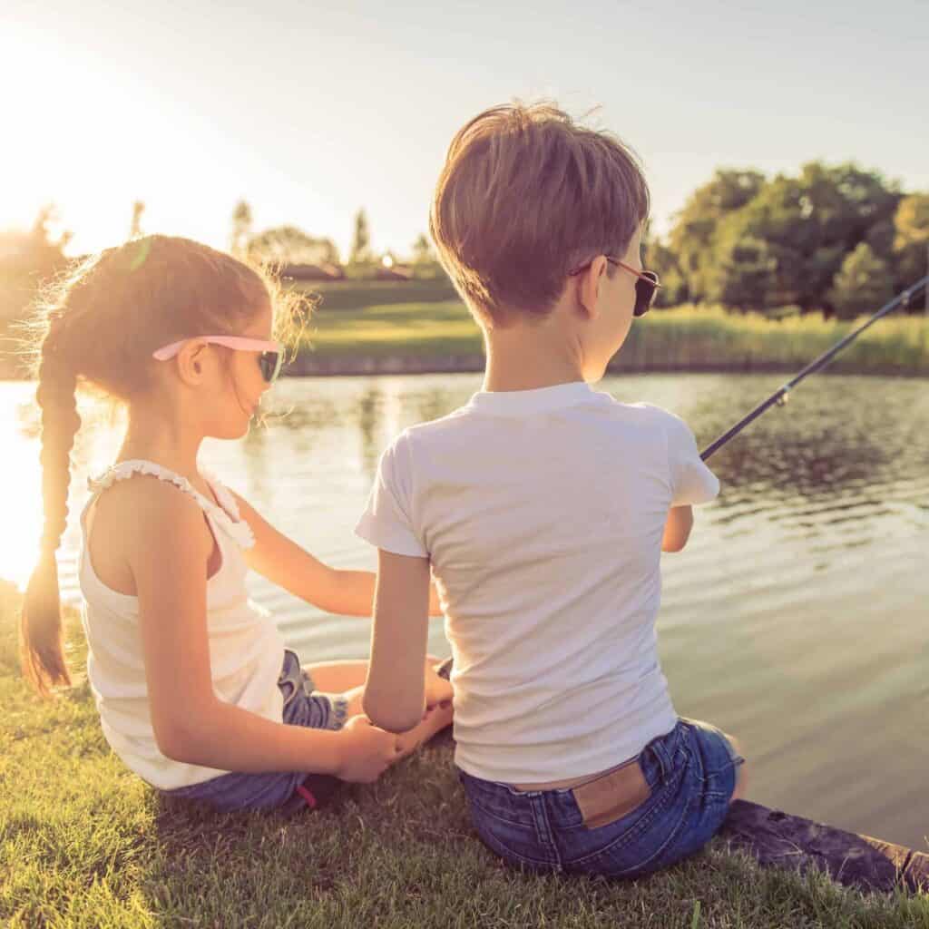 Two children learning how to fish together at the side of a small river.