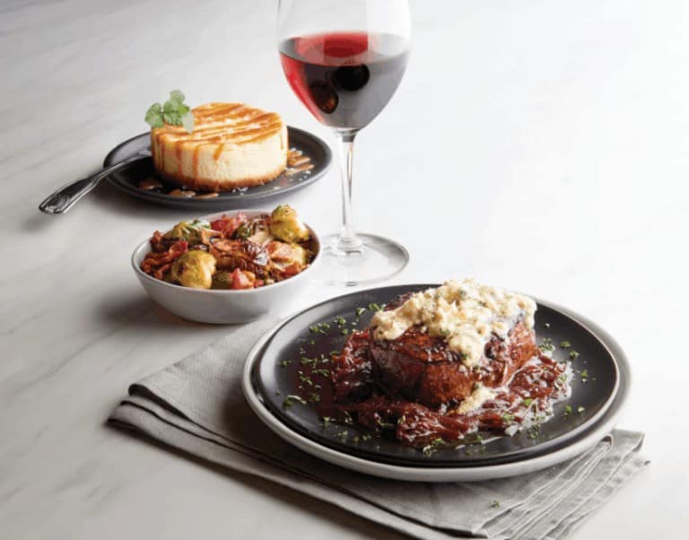 A large steak with toppings and side dishes, including cheesecake and veggies on the side.