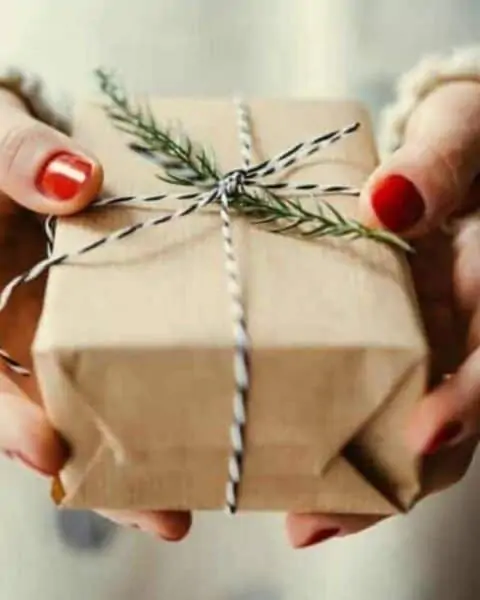 A woman holding a Christmas gift wrapped in brown paper with a green branch from a Christmas tree tied in string.
