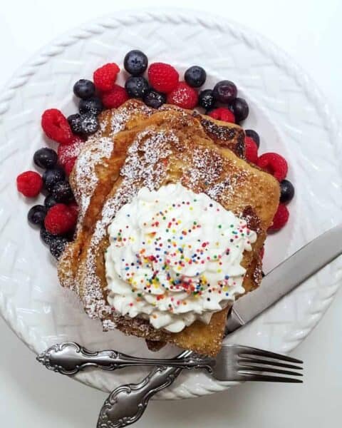 A white plate with French toast piled on top with whip cream and sprinkles. Mixed berries is on the side of the plate.