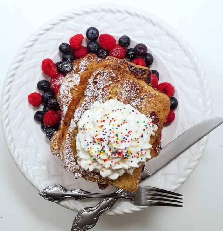 A white plate with French toast piled on top with whip cream and sprinkles. Mixed berries is on the side of the plate.
