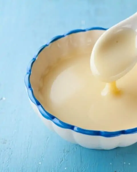 A white pull full of sweetened condensed milk. This is being stirred by a white spoon to mix this recipe together.