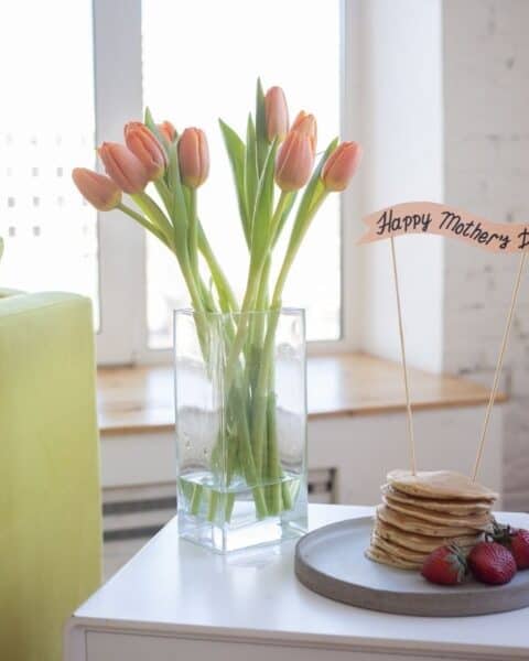 A white tabletop with a stack of pancakes and a vase full of pink tulips.