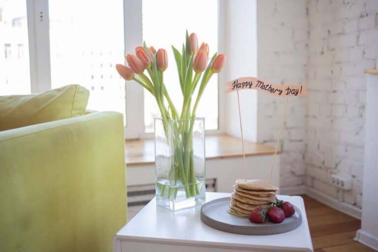 A white tabletop with a stack of pancakes and a vase full of pink tulips.