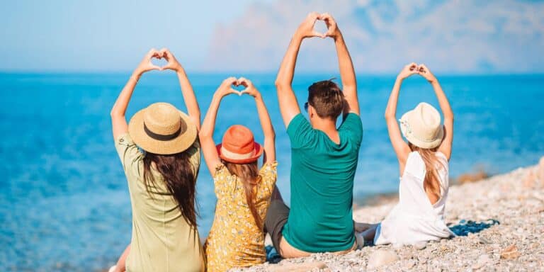A family sitting together on the beach facing the ocean while making heart shapes with their fingers.