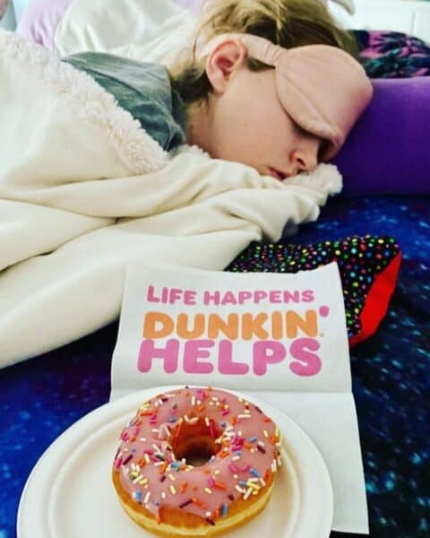 A sleeping woman snuggled with blankets and pillows next to a Dunkin Donuts pink donut.