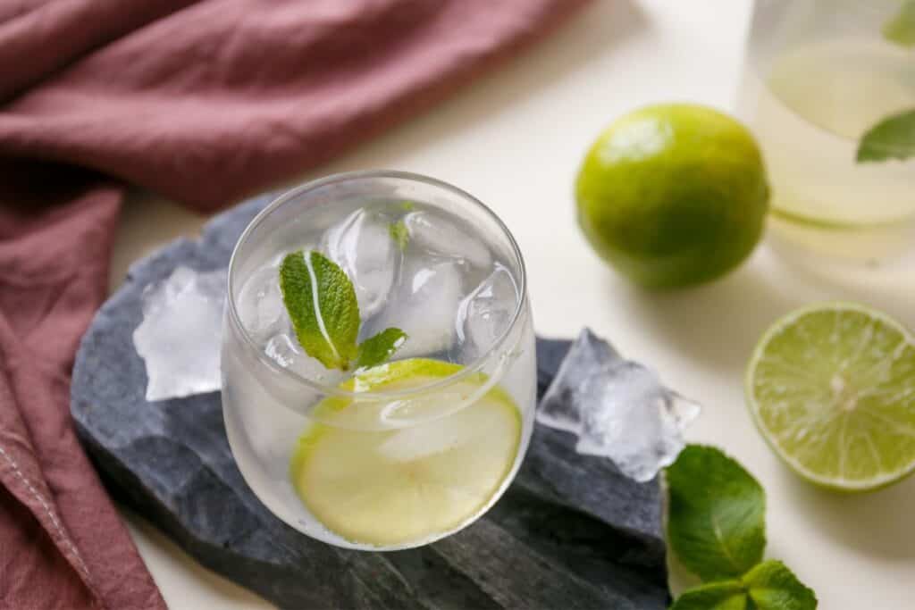 Cup of liquid with a slice of lime and mint. Limes scattered on table top.