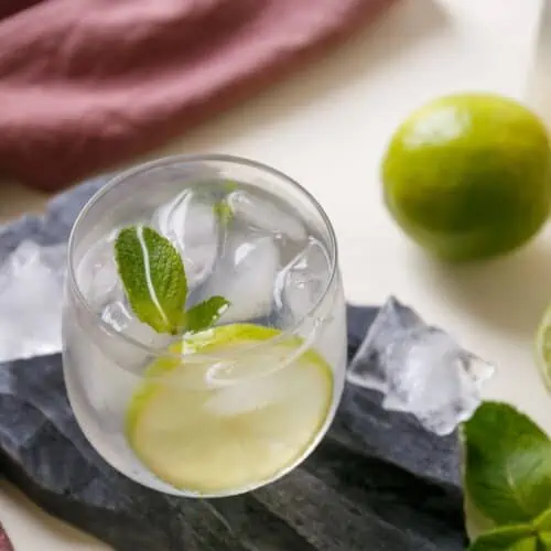 Cup of liquid with a slice of lime and mint. Limes scattered on table top.