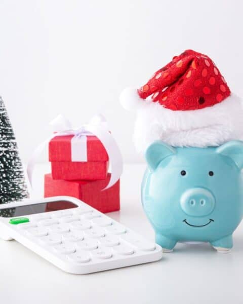 Blue piggy bank with a red Santa hat on its back. The piggy bank is next to a calculator, small tree, and two red presents.