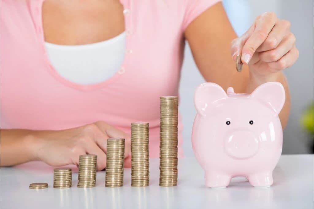 A person is placing a coin in a pink piggy bank next to increasing stacks of coins on a table, demonstrating the right budget method.
