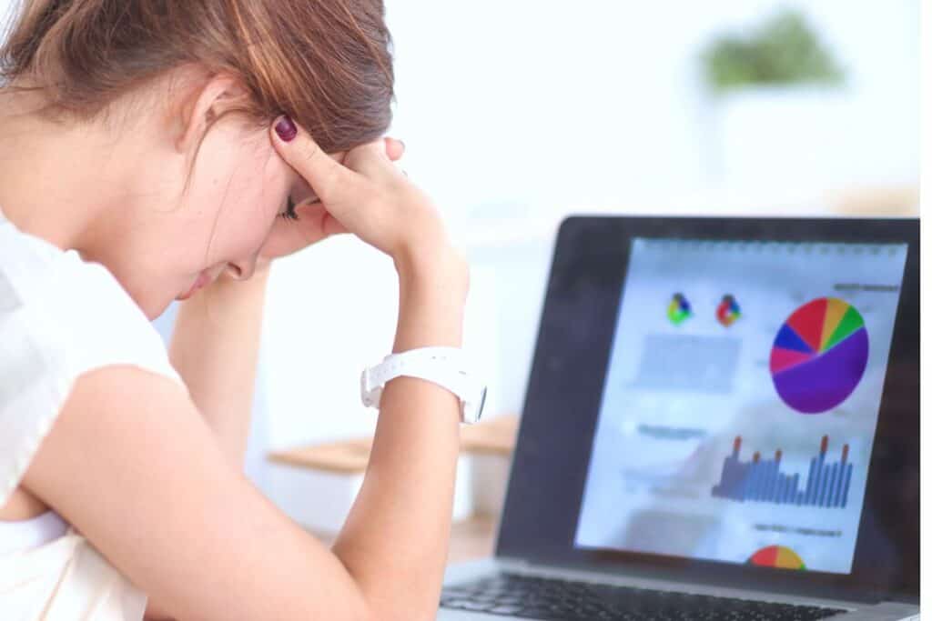 A person with a watch on their wrist sits with their head in their hands, trying to choose the right budget method while looking at a laptop displaying colorful graphs and charts.