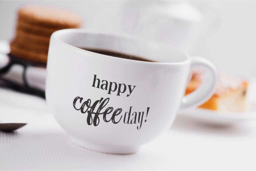 A white coffee cup with "happy Coffee Day!" printed on it, filled with hot coffee, with blurred pastries in the background.