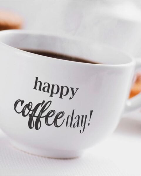 A white coffee cup with "happy Coffee Day!" printed on it, filled with hot coffee, with blurred pastries in the background.