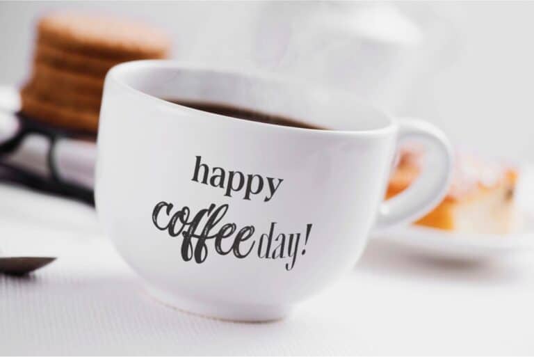 A white coffee cup with "happy Coffee Day!" printed on it, filled with hot coffee, with blurred pastries in the background.