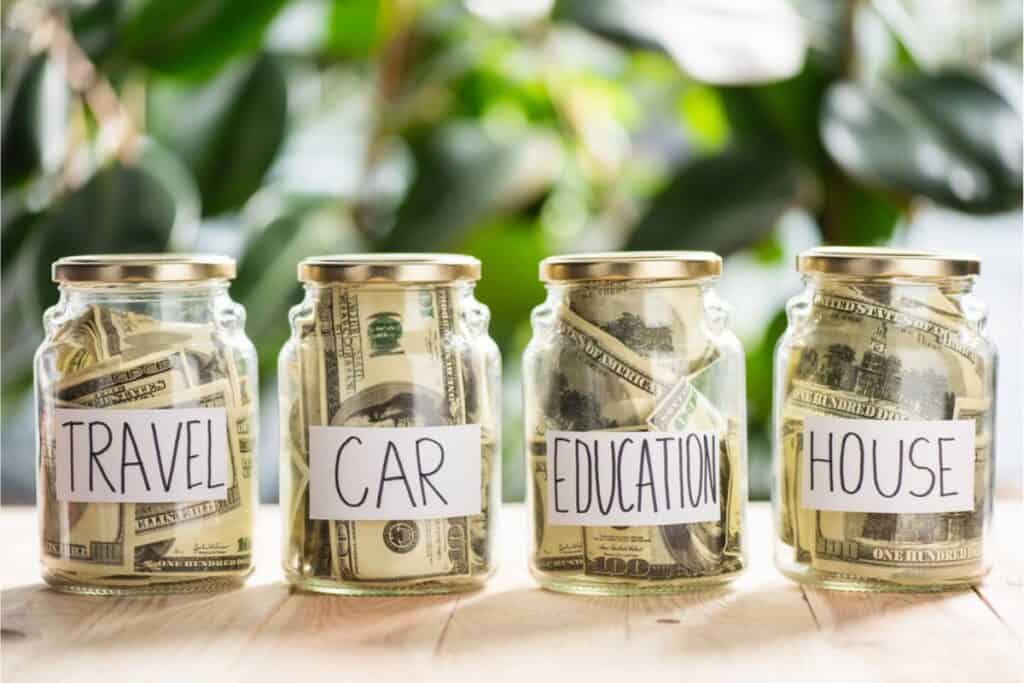 Four glass jars labeled Travel, Car, Education, and House, filled with U.S. dollar bills, are carefully placed on a wooden surface with green leaves in the background. This scene illustrates the Budget Method to help you choose the right financial priorities.