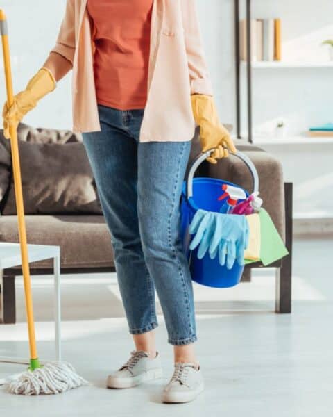 A woman standing in the middle of her living room holding a cleaning bucket in one hand and a mop in the other hand.