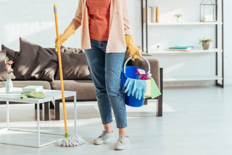 A woman standing in the middle of her living room holding a cleaning bucket in one hand and a mop in the other hand.
