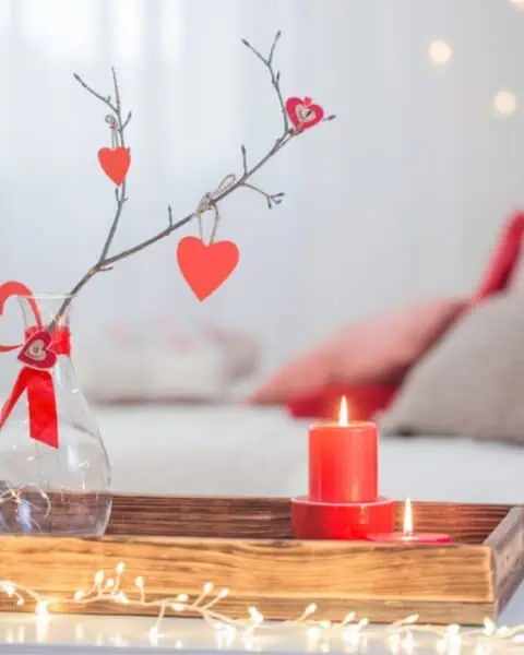A small wooden tray surrounded by lights. There is a small red candle and a white vase with red paper hearts hanging off of the branch in the vase.