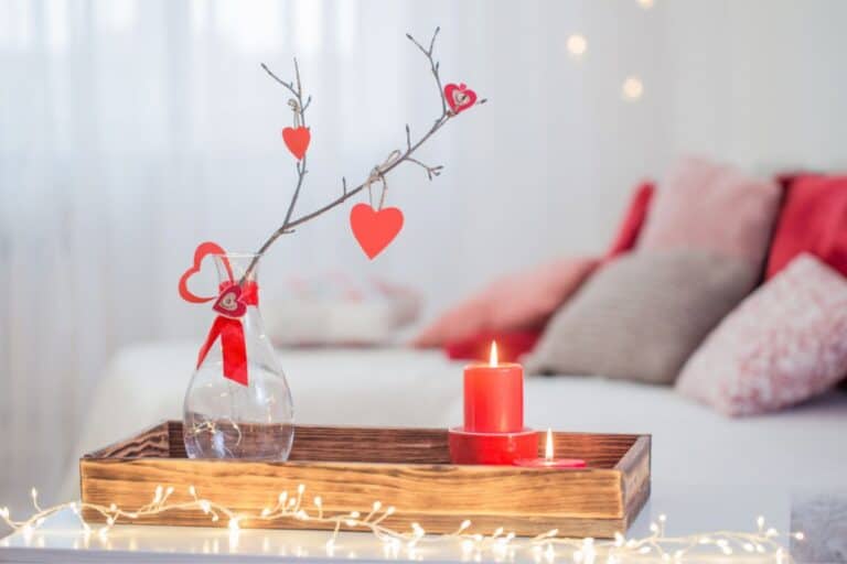A small wooden tray surrounded by lights. There is a small red candle and a white vase with red paper hearts hanging off of the branch in the vase.