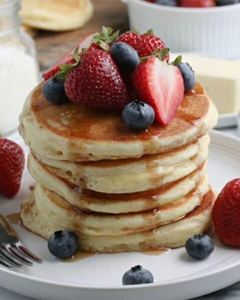 A white plate holding five pancakes. The pancakes are garnished with fresh berries and pancake syrup.