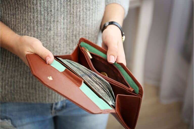 A woman opening up her brown feminine wallet and she has a stack of cash and coins inside.