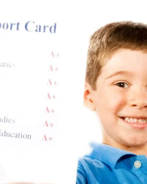 A young boy with a happy smile holding up a report card with all A+'s.