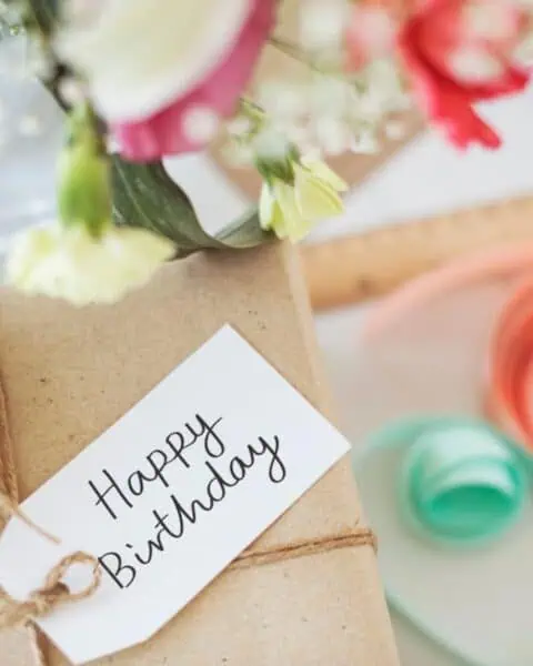 A brown paper package with a "Happy Birthday" tag, surrounded by flowers and colored ribbons, promises delightful birthday freebies within its charming wrap.