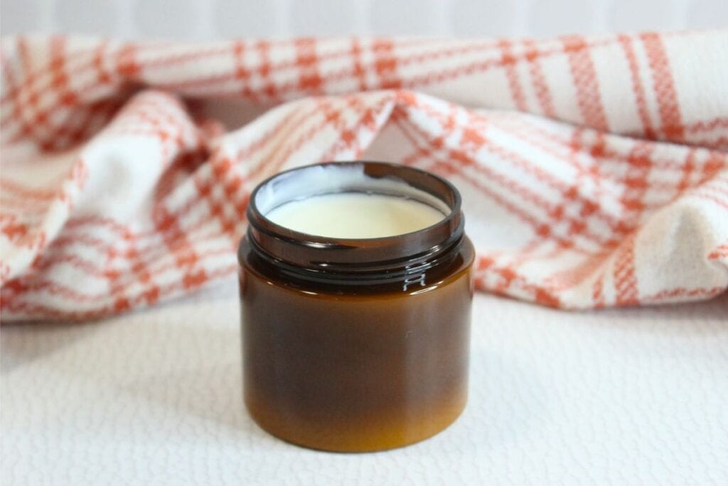 A small, open brown jar of natural sunscreen filled with white cream sits in front of a red and white checkered cloth.