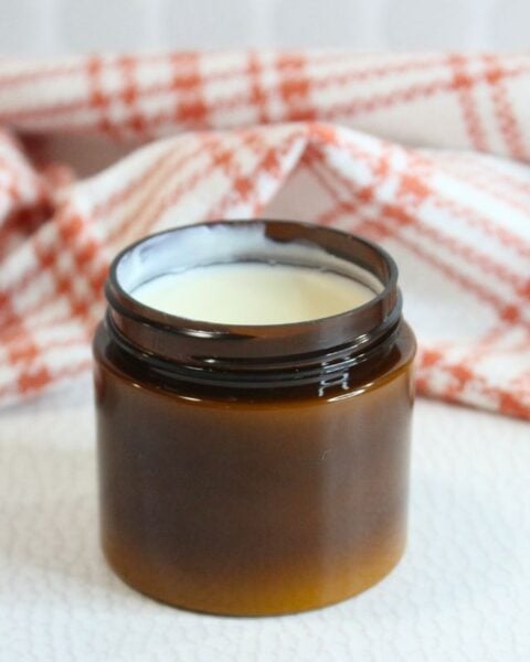 A small, open brown jar of natural sunscreen filled with white cream sits in front of a red and white checkered cloth.