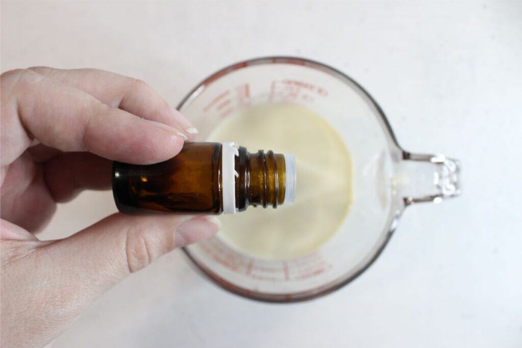 A hand holds a small amber bottle over a glass measuring cup containing a creamy liquid, as if about to add drops to the natural sunscreen mixture.
