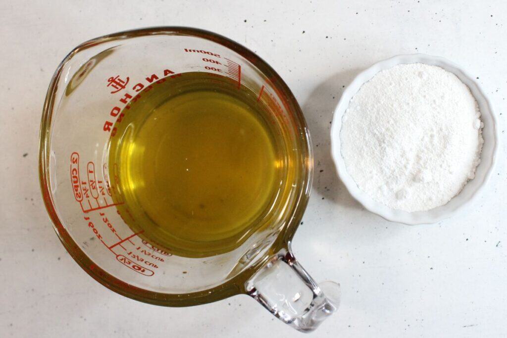 A glass measuring cup filled with oil sits next to a small white bowl of white powder on a light surface—perfect for crafting your own natural sunscreen at home.