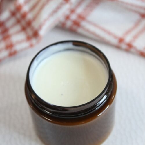 An open brown glass jar filled with creamy white natural sunscreen sits on a light surface, with a red and white checkered cloth in the background.