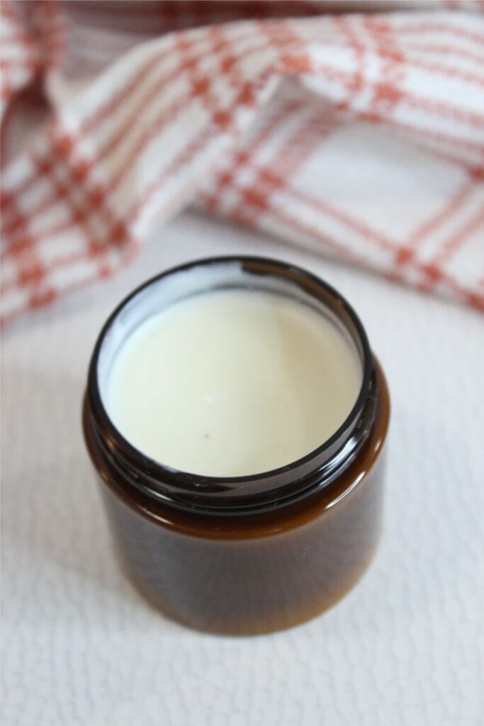An open brown glass jar filled with creamy white natural sunscreen sits on a light surface, with a red and white checkered cloth in the background.