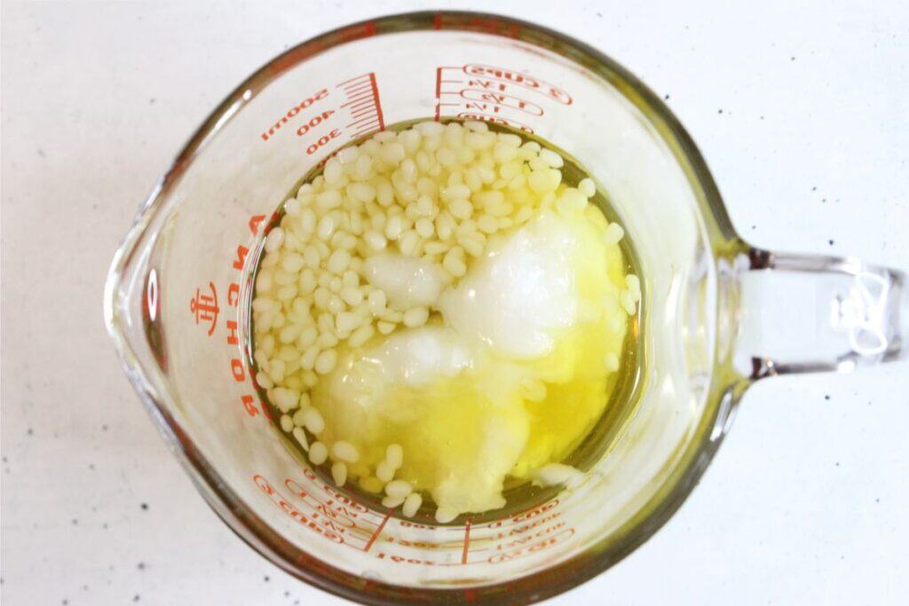 A glass measuring cup containing solid white pellets, white clumps, and yellow liquid—common ingredients when making a natural sunscreen—viewed from above on a white surface.