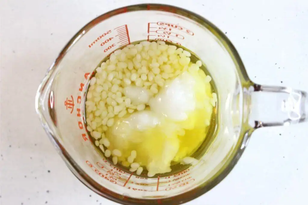 A glass measuring cup containing solid white pellets, white clumps, and yellow liquid—common ingredients when making a natural sunscreen—viewed from above on a white surface.