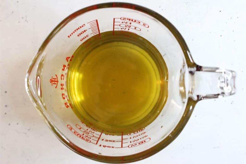 A glass measuring cup filled with a yellow liquid, possibly a homemade natural sunscreen, viewed from above on a white surface.