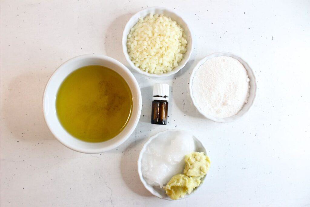 Five ingredients in bowls—liquid oil, beeswax pellets, a small bottle, white powder, and two types of butter—are arranged on a white surface, ready to be blended into a nourishing natural sunscreen.