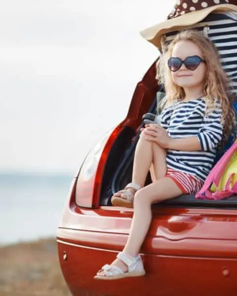 A little girl sitting in a car trunk, packed with luggage for a family road trip.