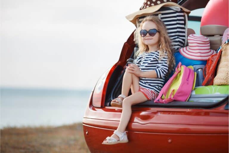 A little girl sitting in a car trunk, packed with luggage for a family road trip.