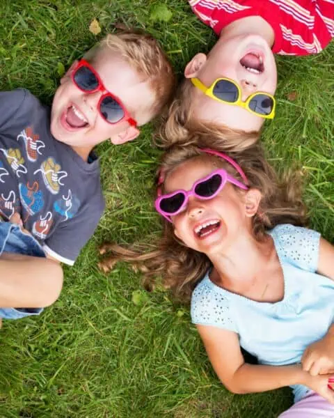Three children enjoying summer by laying on the grass, wearing sunglasses.