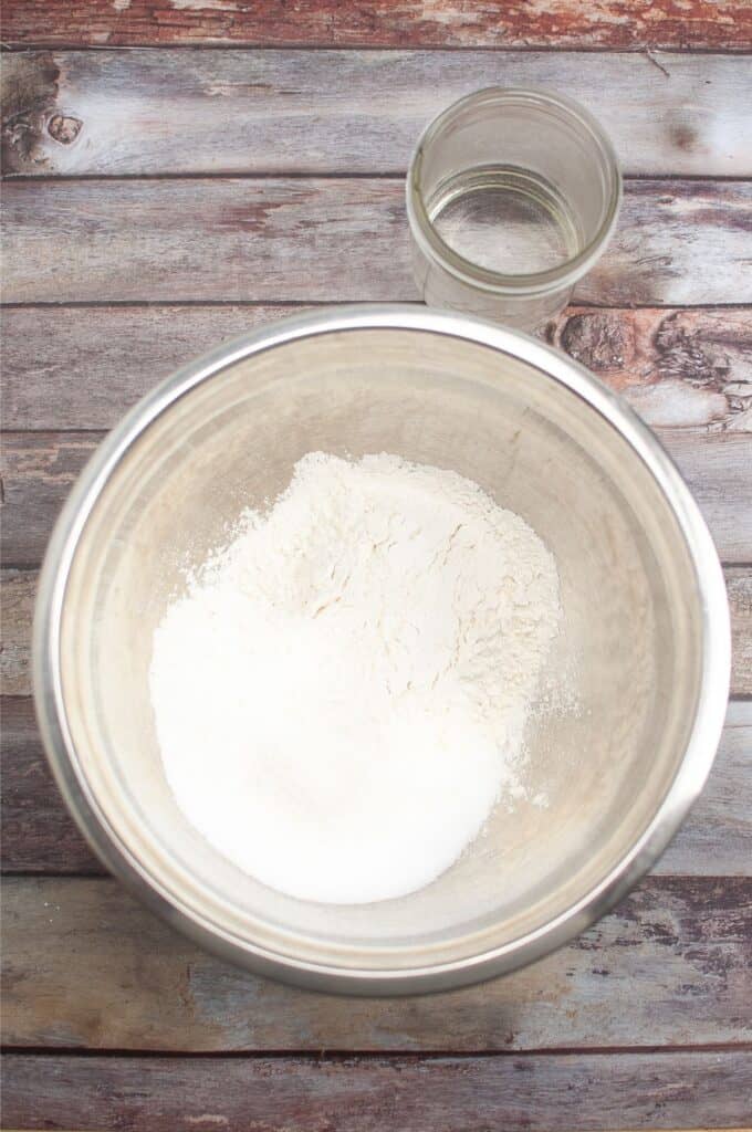 A bowl of flour next to a glass on a wooden table, used for making Salt Dough Ornaments.