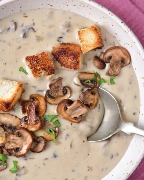A bowl of cream of mushroom soup with croutons and a spoon.